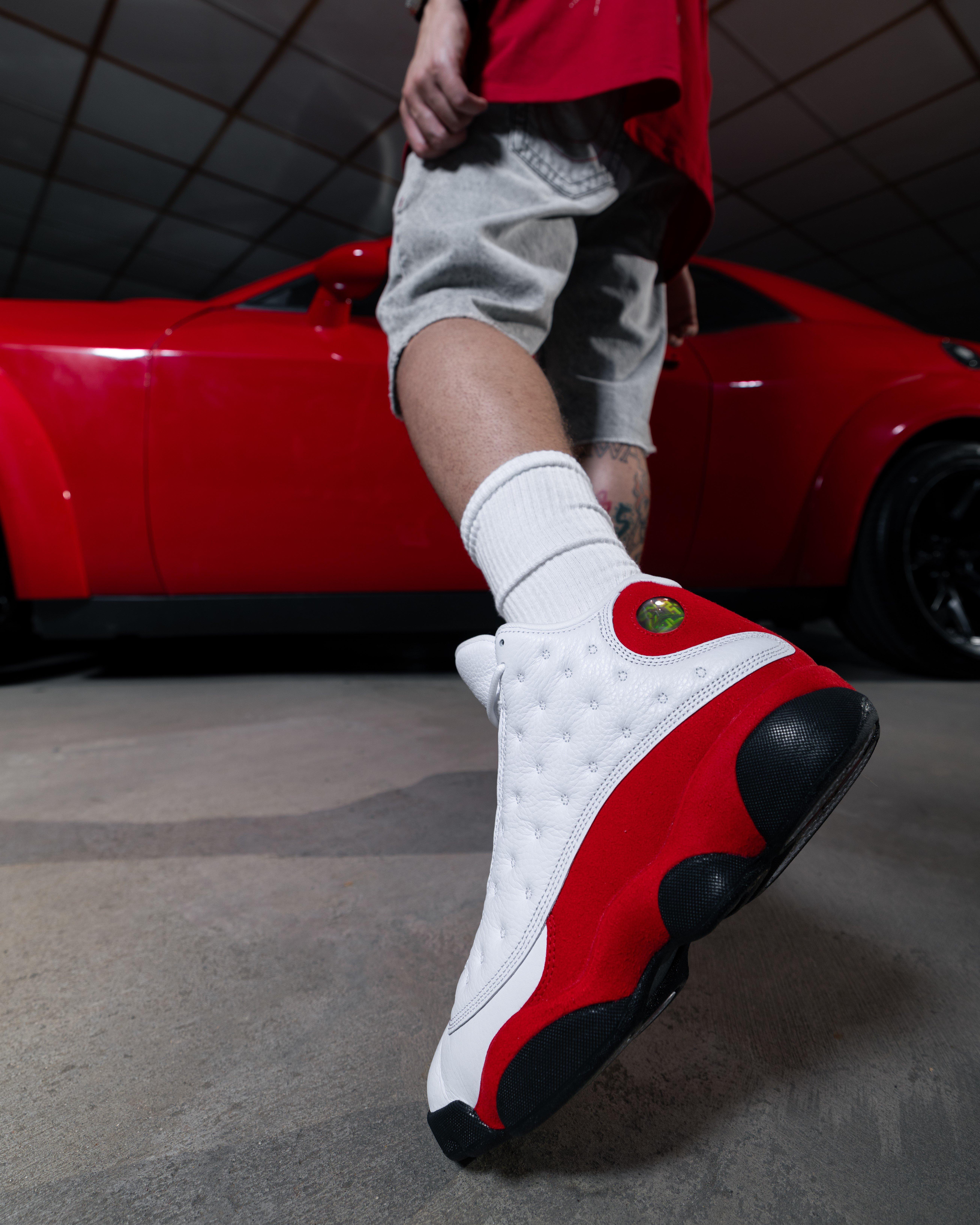Model walking in white and red Air Jordan 13 sneakers in front of a red sports car, showing the side and outsole design.